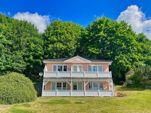 Une maison à deux étages dotée d’un balcon, entourée d’une végétation luxuriante et d’arbres.