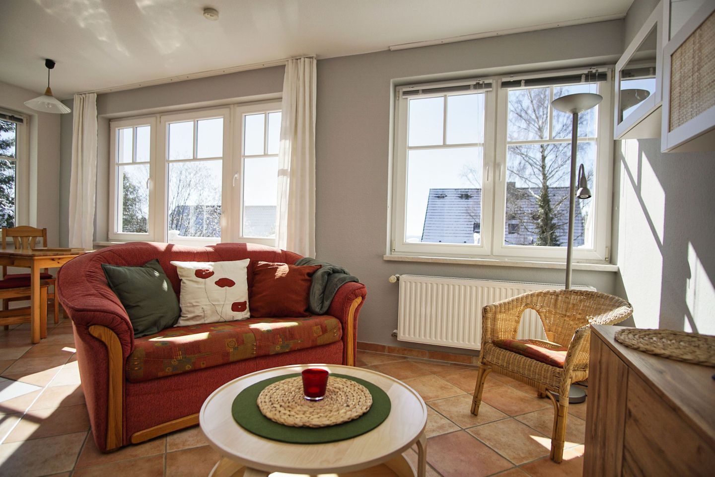 Ein Wohnzimmer mit einem roten Sofa, einem Korbsessel, einem kleinen Tisch mit einer roten Tasse und großen Fenstern mit Blick auf Gebäude und Bäume.