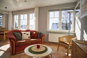Un salon avec un canapé rouge, un fauteuil en osier, une petite table sur laquelle se trouve une tasse rouge, et de grandes fenêtres offrant une vue sur des immeubles et des arbres.