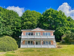 Une maison à deux étages dotée d’un balcon, entourée d’une végétation luxuriante et d’arbres.