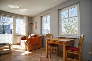 A cozy living room with a sofa, a wooden dining table, and chairs. There are large windows providing natural light and views of trees outside.