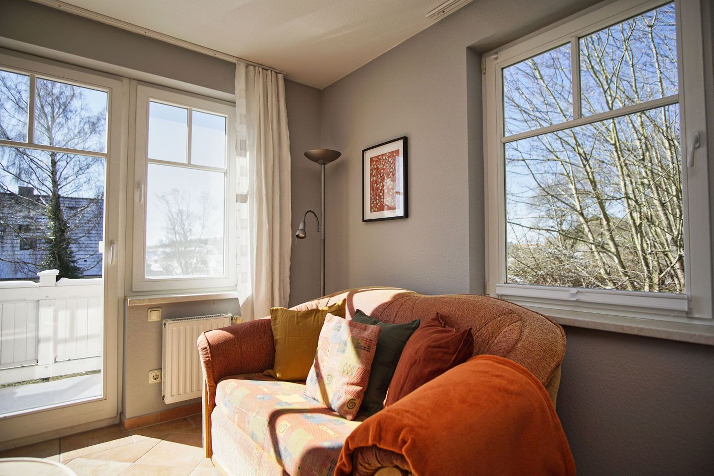 Ein gemütliches Wohnzimmer mit einer Couch, einer Heizung und großen Fenstern mit Blick auf Bäume und ein Gebäude.