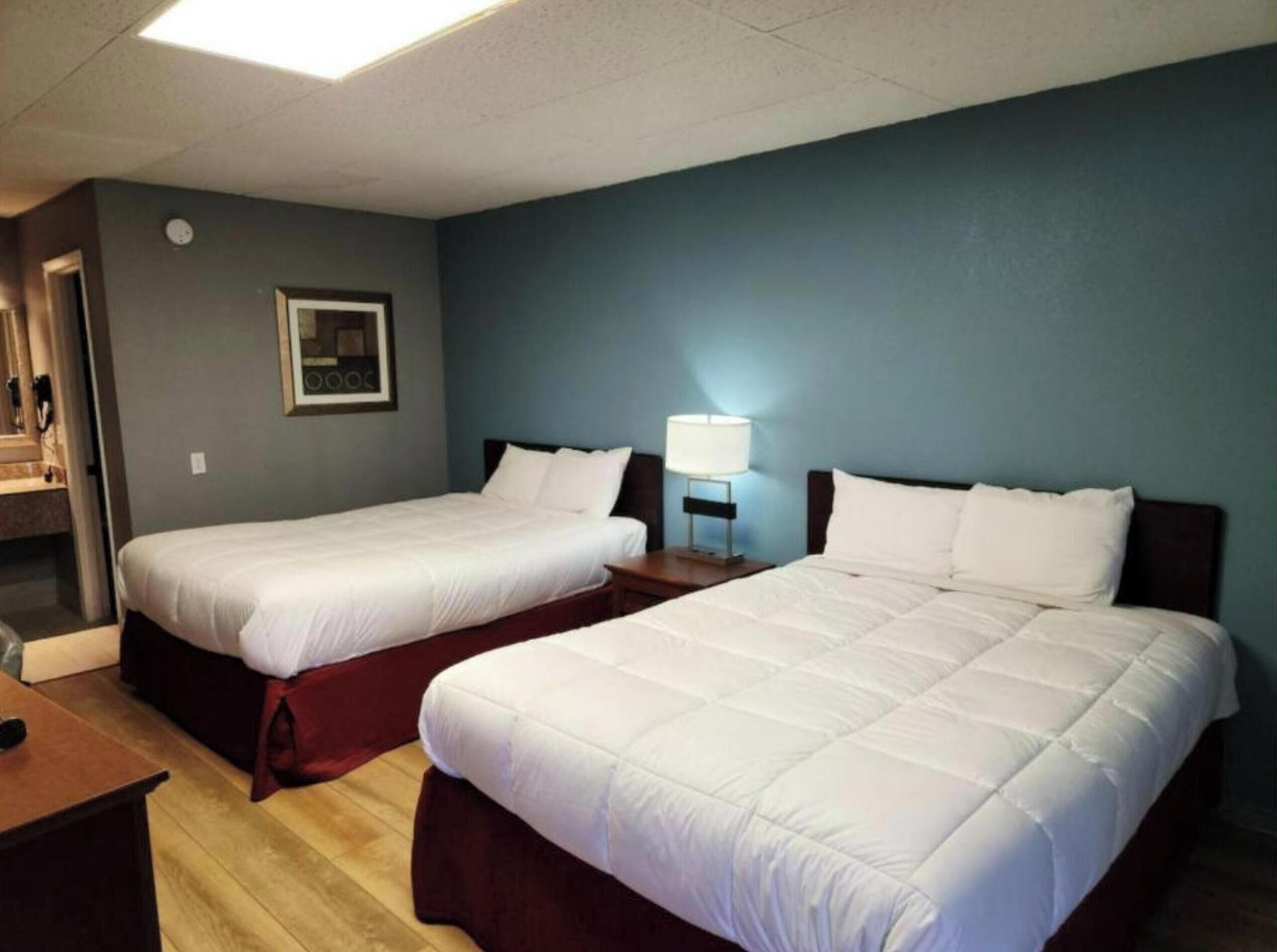 Habitación de hotel con dos camas, una mesita de noche con lámpara, un cuadro enmarcado en la pared y suelo de madera.