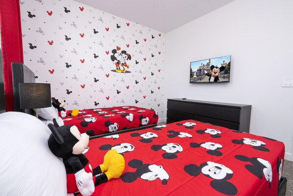 A room with two beds, each covered with a Mickey Mouse themed bedspread. There is a Mickey Mouse plush toy on one bed.