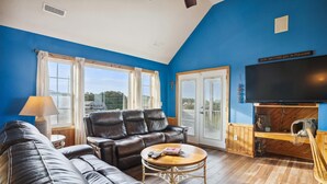 A living room with a blue wall, a leather sofa, a wooden coffee table, and a television. - 7130- Sunset Breeze - Soundfront Waves | Pool & Sunset Views (Waves)