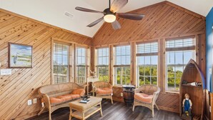 A cozy room with wooden walls, large windows, wicker furniture, and a ceiling fan. - 7130- Sunset Breeze - Soundfront Waves | Pool & Sunset Views (Waves)