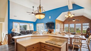 A spacious kitchen with a central island, wooden cabinets, and a large window providing a view of the outdoors. - 7130- Sunset Breeze - Soundfront Waves | Pool & Sunset Views (Waves)