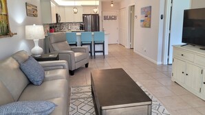 A modern living room with a sofa, coffee table, and TV. There is a kitchen in the background with a refrigerator and cabinets. - OV 914 Partial Course View Condo - Paradise Rental in Ocean Village (Fort Pierce)