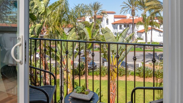 A balcony with two chairs and a small table, a glass door, and a view of palm trees and buildings. - 645 Fresh Modern Condo Inside Resort Gates - Free Wifi - Walk to Spa (Carlsbad)