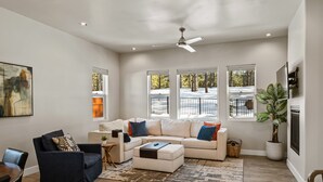 A spacious living room with a sectional sofa, a dark armchair, and a coffee table. There is a ceiling fan, large windows with a view of trees, and a potted plant. - Pinnacle Pines | Forest Views | Modern Home with EV Charger | Direct Trail Access (Flagstaff)