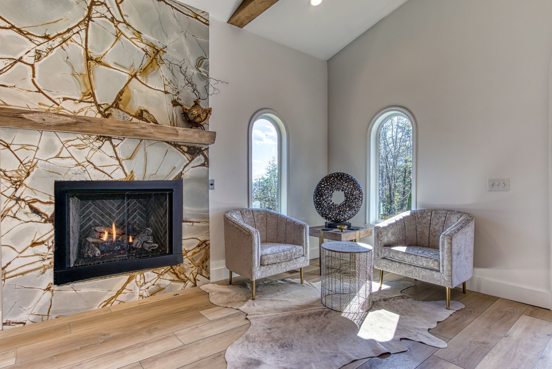 A modern living room with a fireplace, two armchairs, a round table, and a large window with a view of trees.