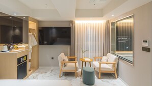 Une chambre d’hôtel moderne équipée d’un lit, d’un bureau avec un ordinateur, d’une télévision, de deux fauteuils, d’une petite table et d’une grande fenêtre avec des rideaux.