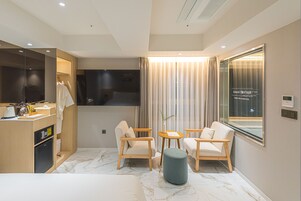 Una habitación de hotel moderna con una cama, un escritorio con computadora, televisor, dos sillas, una mesita y un ventanal con cortinas.