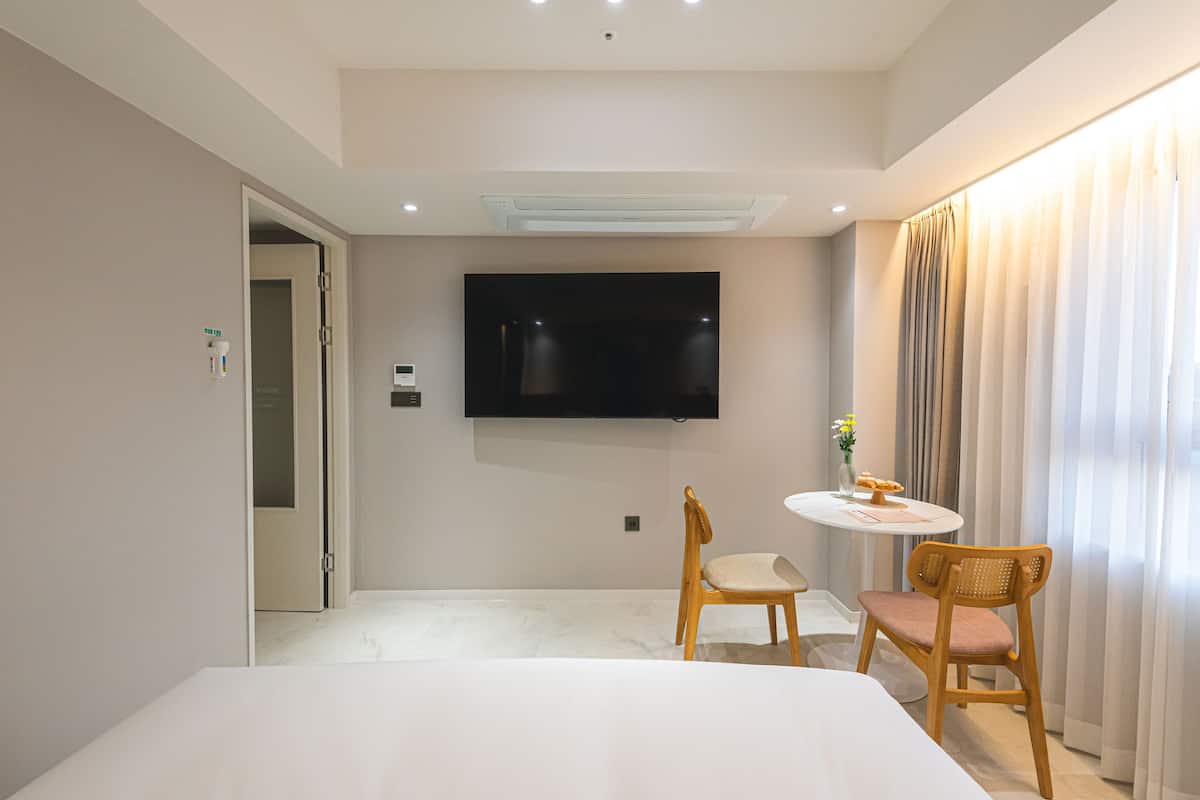 Ein Hotelzimmer mit Bett, Wand-Fernseher, einem kleinen Tisch mit Blumenvase und einem Sessel.