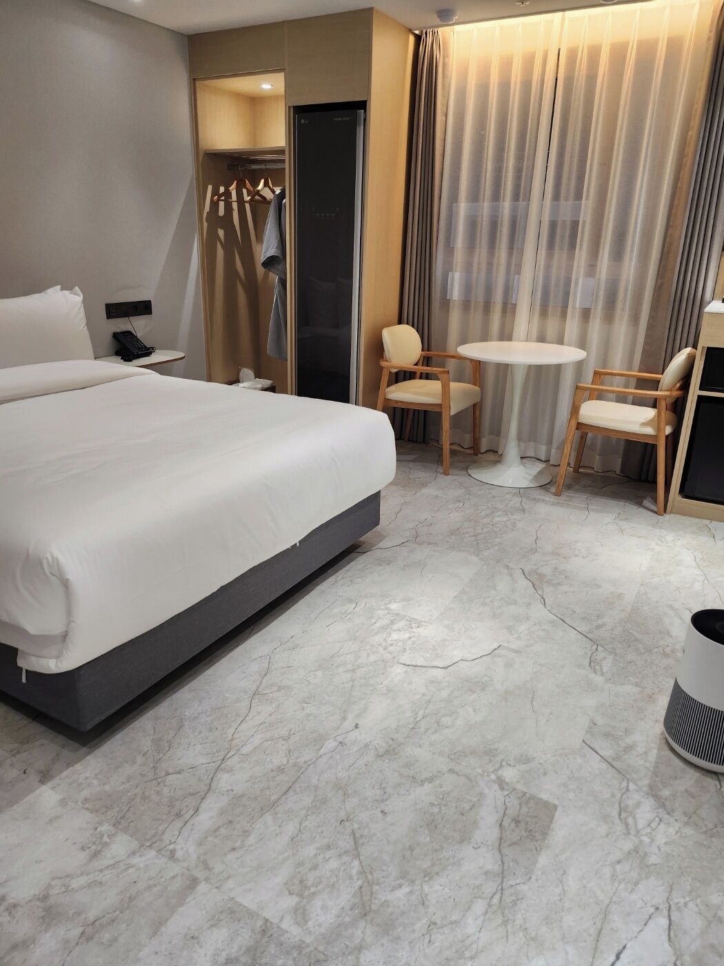 A modern hotel room with a large bed, a small round table, two chairs, a wardrobe, and a bathroom door.