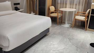A modern hotel room with a large bed, a small round table, two chairs, a wardrobe, and a bathroom door.