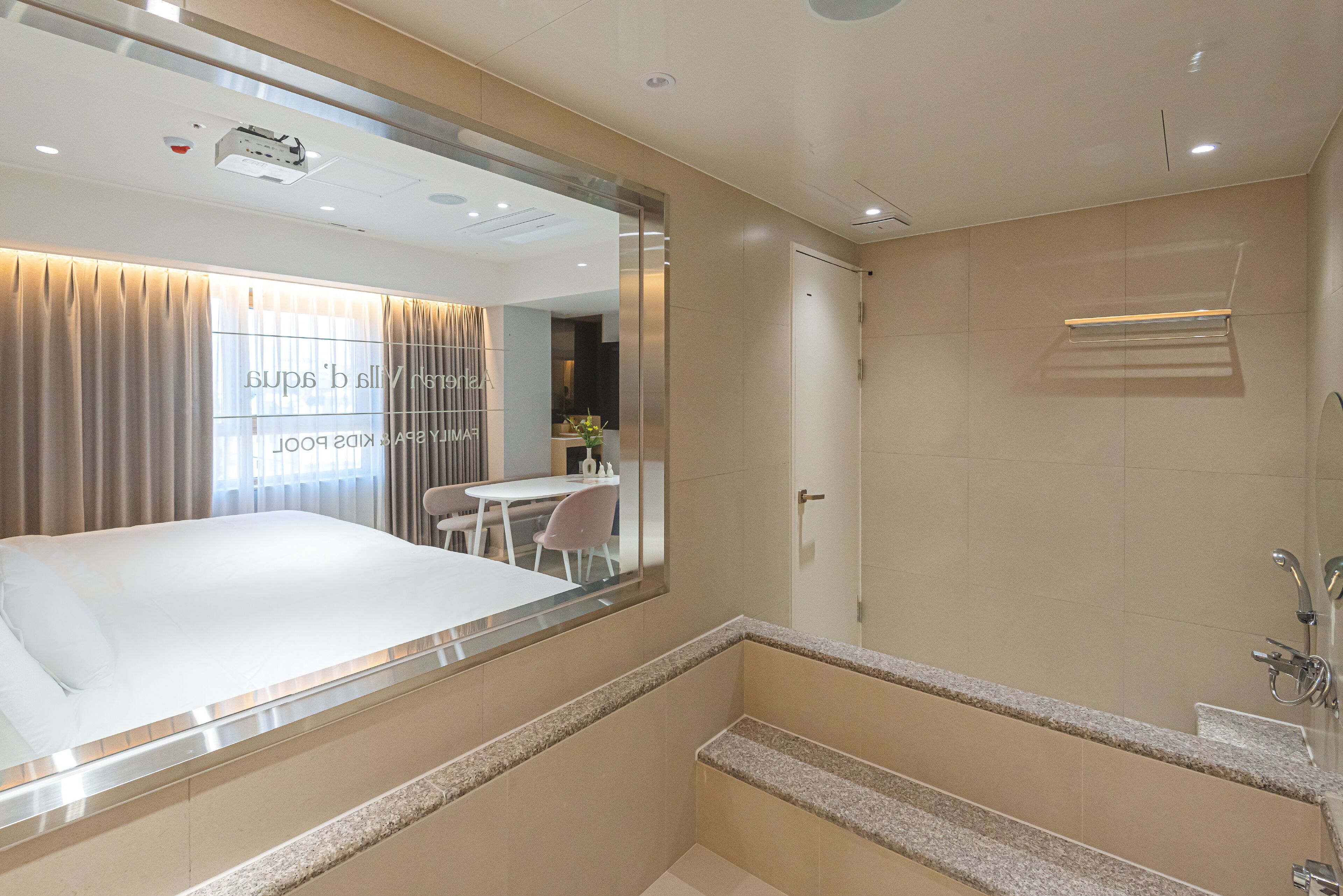 A modern hotel room with a large bed, a bathroom with a bathtub, and a dining area visible through a glass partition.