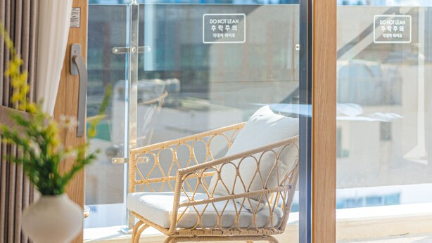 Une chambre moderne avec une porte en verre, un fauteuil en osier, une table sur laquelle se trouvent un gâteau et une tasse de café, et une vue sur de hauts immeubles.