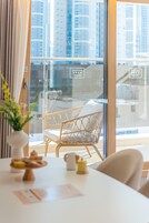 Una habitación moderna con una puerta de vidrio, una silla de mimbre, una mesa con un pastel y una taza de café, y vistas de edificios altos.