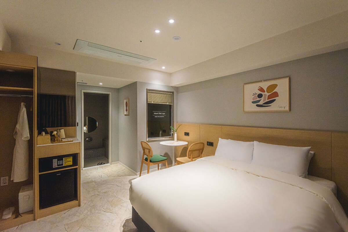Ein Hotelzimmer mit einem großen Bett, einem Kleiderschrank, einem kleinen Tisch mit Stühlen und einem durchgehenden Türspalte sichtbaren Badezimmer.