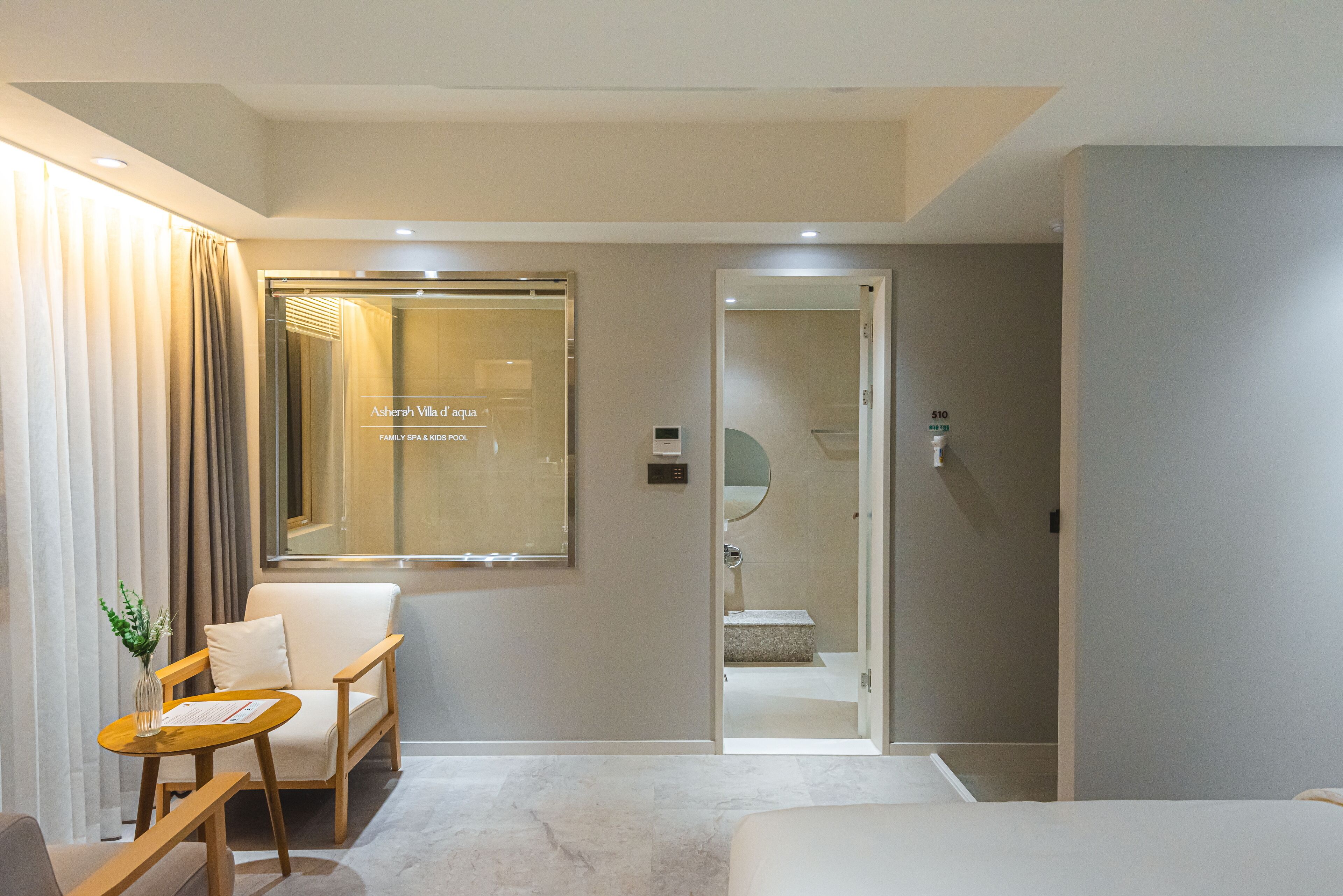 A modern hotel room with a bed, a seating area with a chair and a small table, a large window with curtains, and a visible bathroom with a shower and a mirror.