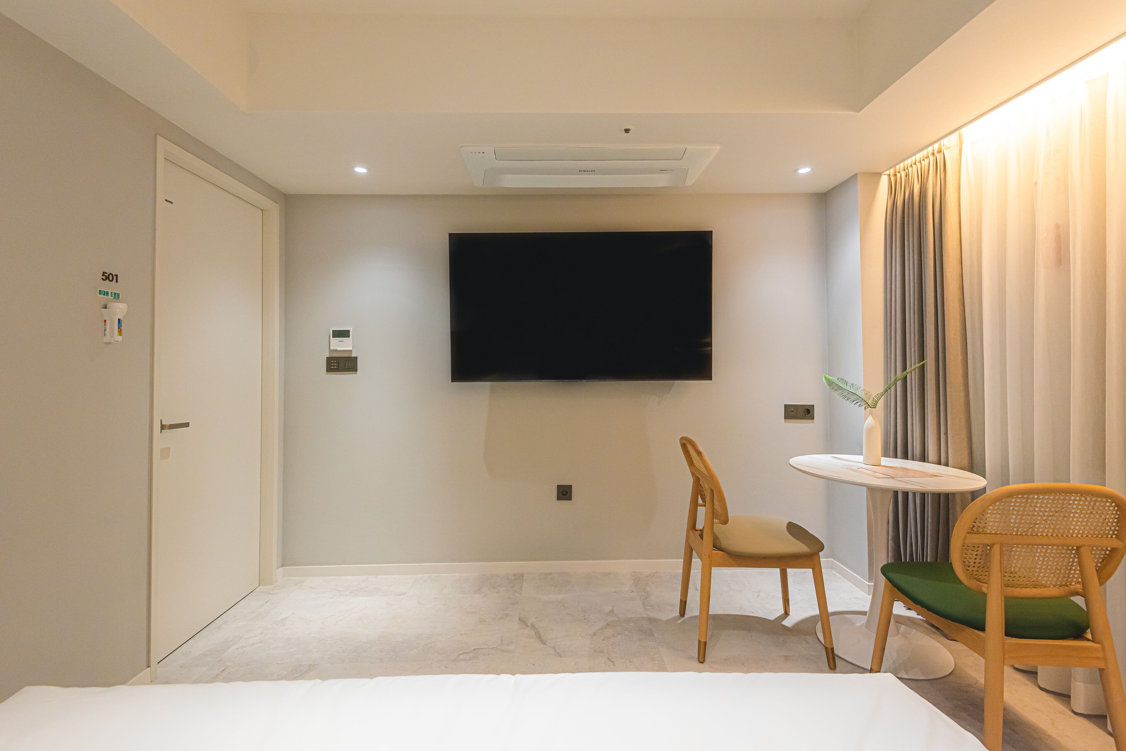 Une chambre d’hôtel équipée d’un téléviseur à écran plat fixé au mur, d’un lit, d’une chaise, d’une petite table et d’une porte portant le numéro « 501 ».