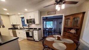 Une cuisine avec des meubles blancs, un fourneau et un ventilateur de plafond. On y trouve également un coin repas avec une table et des chaises.