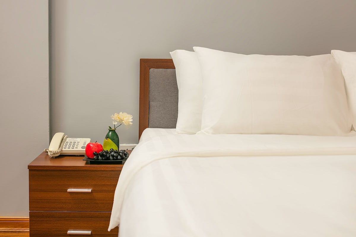 A neatly made bed with white linens, a wooden nightstand with a telephone and fruit, and a vase with flowers.