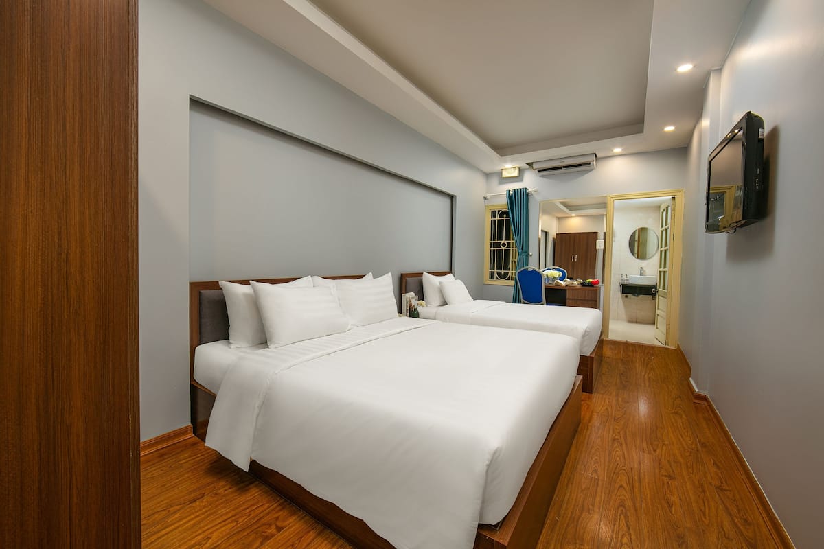 A hotel room with two beds, wooden flooring, a TV mounted on the wall, and a bathroom visible through an open door.