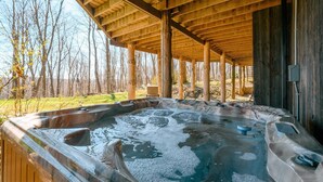 Une cabane en bois avec un jacuzzi situé sous une zone abritée, entourée d’arbres.