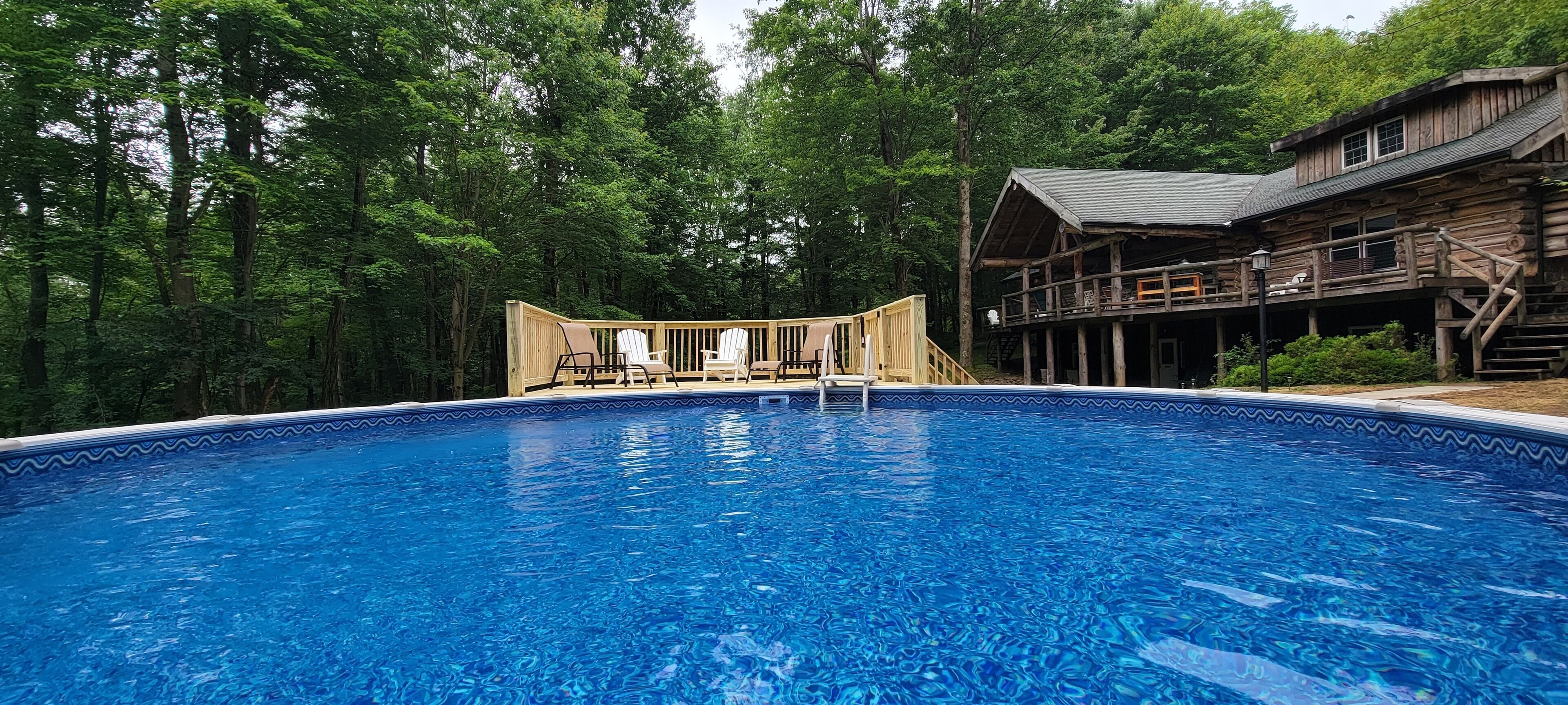 Une piscine avec une terrasse en bois et des chaises, entourée d’arbres.