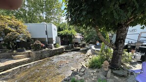 Un petit riu amb un pont de pedra, envoltat de vegetació i una caravana blanca.