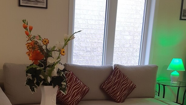 A living room with a beige sofa, a glass coffee table, a vase with flowers, a lamp, and a window with blinds.