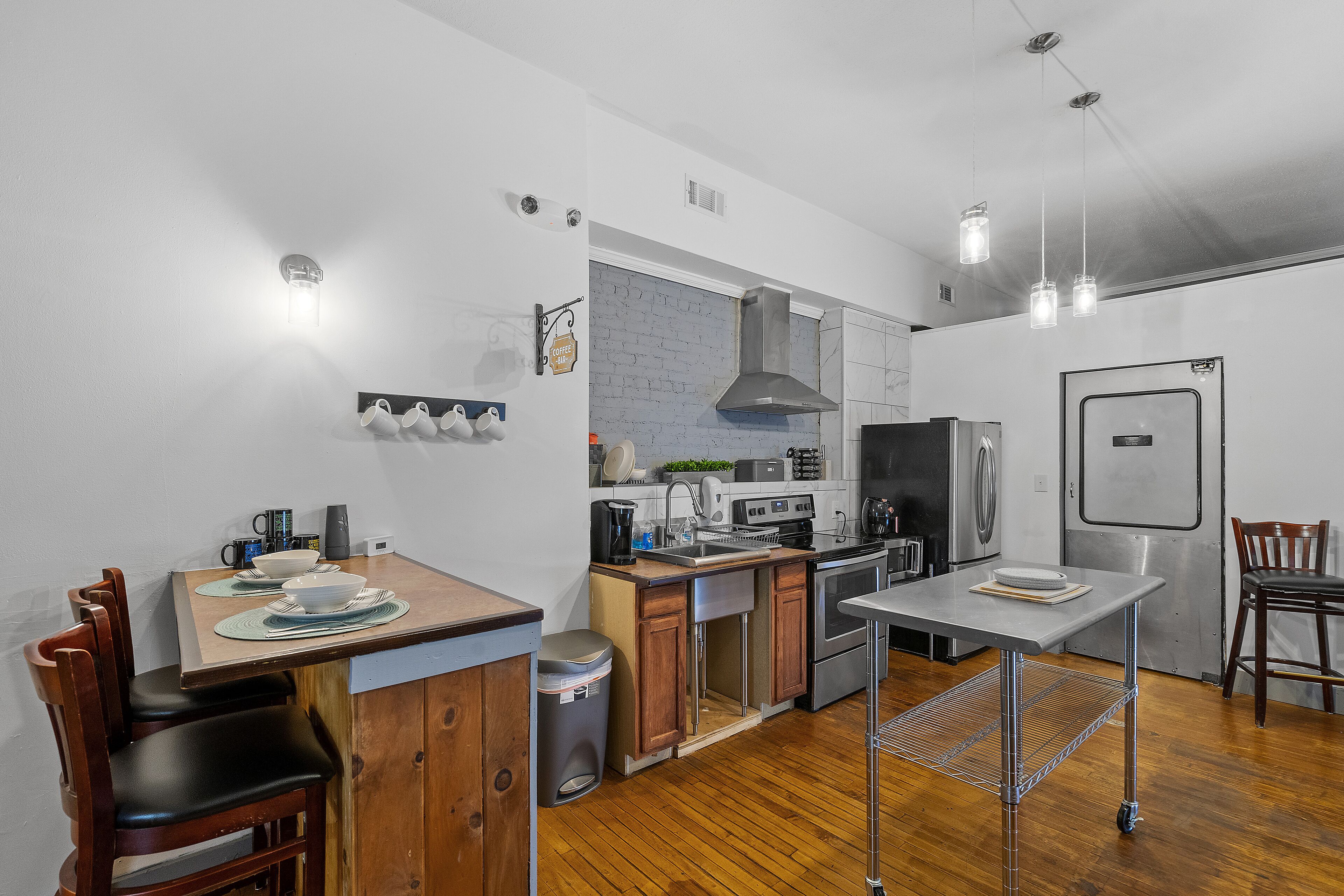 A compact kitchen with wooden cabinets, a stainless steel sink, and a built-in oven. There is a dining area with a table and chairs, and a trash can is visible.