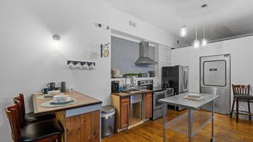 A compact kitchen with wooden cabinets, a stainless steel sink, and a built-in oven. There is a dining area with a table and chairs, and a trash can is visible.