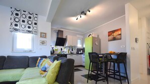 A modern living room with a sofa, a dining area with a table and chairs, a kitchen with a fridge and a TV, and a window with blinds. - Apartment Diva near the beach (Njivice)