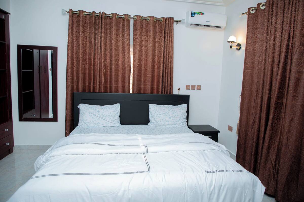 A neatly made bed with white bedding, a dark headboard, and brown curtains.