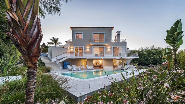 Una casa de dos pisos con piscina, rodeada de exuberante follaje y plantas con flores.