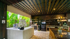 A modern kitchen with a central island, a dining area with a table and chairs, and a view of lush greenery outside. - Tanaga Coliving (Seminyak)