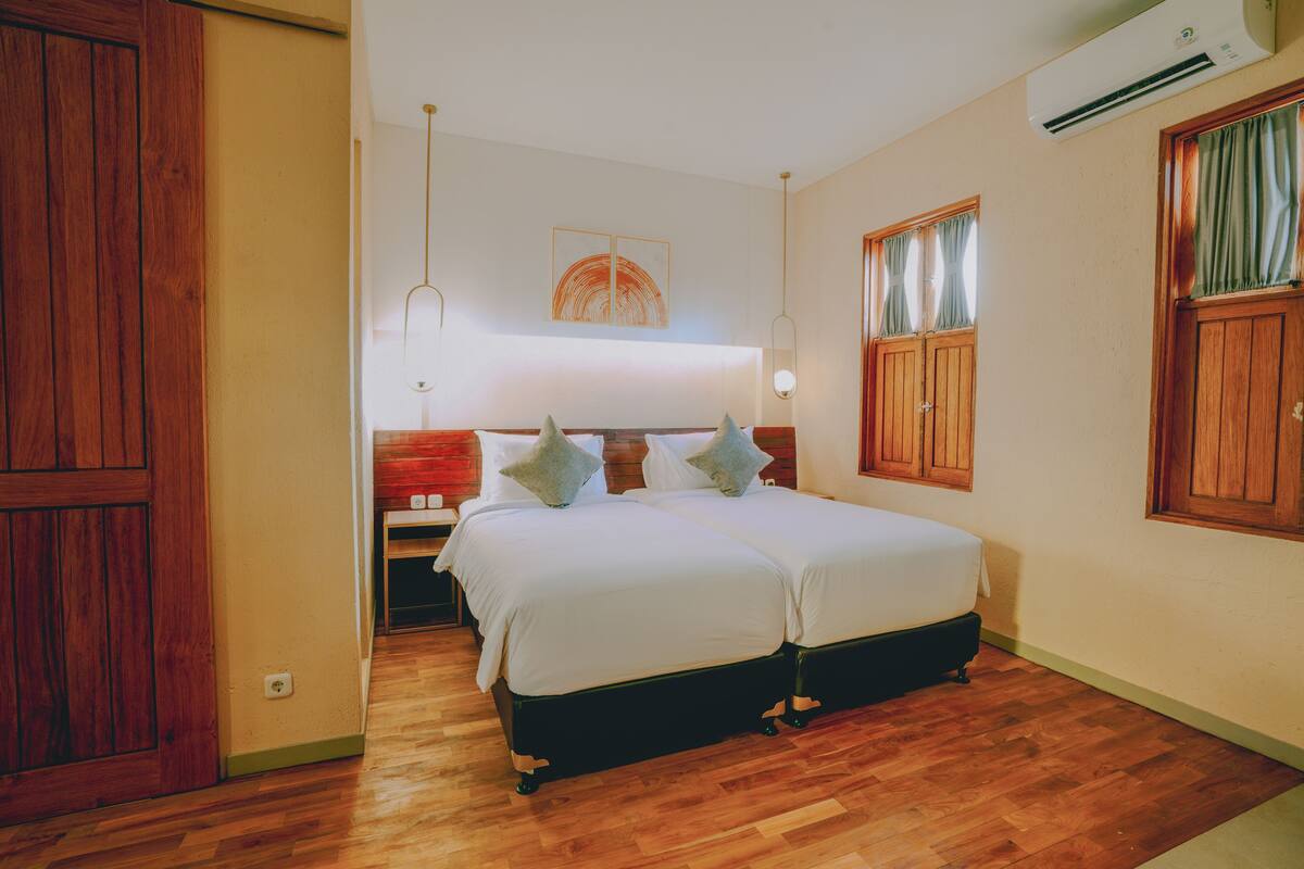 Quarto com duas camas, soalho de madeira, uma porta grande de madeira e um aparelho de ar condicionado.