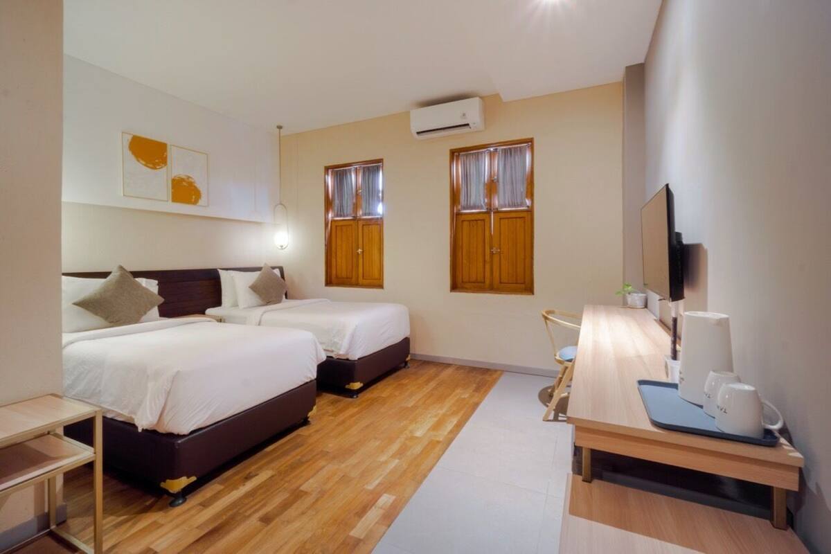 Quarto de hotel com duas camas, chão de madeira, escrivaninha com cadeira, televisão e ar condicionado.