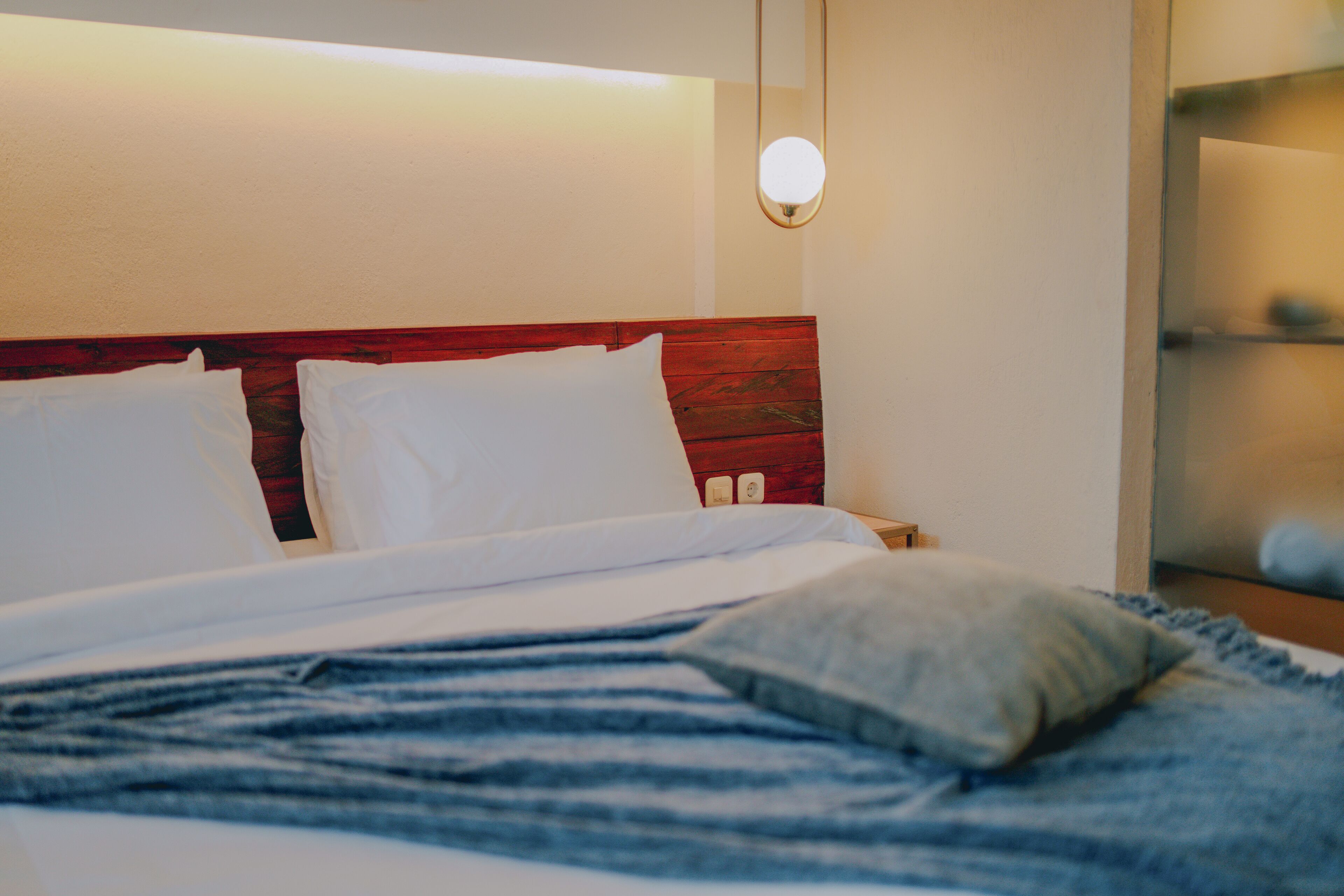Una cama bien hecha con sábanas blancas y una manta azul a rayas, un cabecero de madera y una mesita de noche con una lámpara.