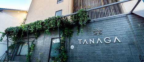 A building with a sign reading "TANAGA" and a modern design with a brick facade and a metal railing.