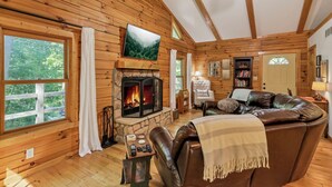 A cozy log cabin interior with a stone fireplace, leather sofa, and a television mounted on the wall. - Minutes to Caves - 2 KINGS. Hot Tub. Games. Firepit. Close to Everything (Logan)