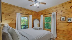 A wooden-paneled room with a bed, two windows with curtains, a ceiling fan, and framed pictures on the wall. - Minutes to Caves - 2 KINGS. Hot Tub. Games. Firepit. Close to Everything (Logan)