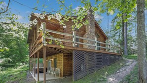 A wooden cabin with a covered porch, stone chimney, and a view of the surrounding forest. - Minutes to Caves - 2 KINGS. Hot Tub. Games. Firepit. Close to Everything (Logan)