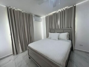 A modern bedroom with a large bed, grey curtains, and an air conditioning unit on the wall. - Punta Cana - Villa El Encanto (Punta Cana)
