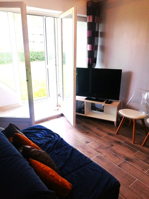 A room with a bed, a TV on a stand, a chair, and a sliding glass door leading to an outdoor area. - Close to amenities with good exposure (Cabourg)
