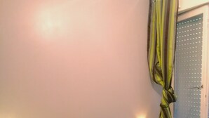 Un dormitorio con cama, dos almohadas, una mesita de noche con lámpara, una ventana con cortinas y una puerta con manija.