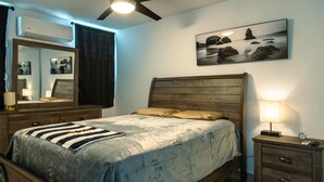 A bedroom with a wooden bed, a nightstand with a lamp, a ceiling fan, a wall-mounted air conditioner, and a framed picture of rocks.